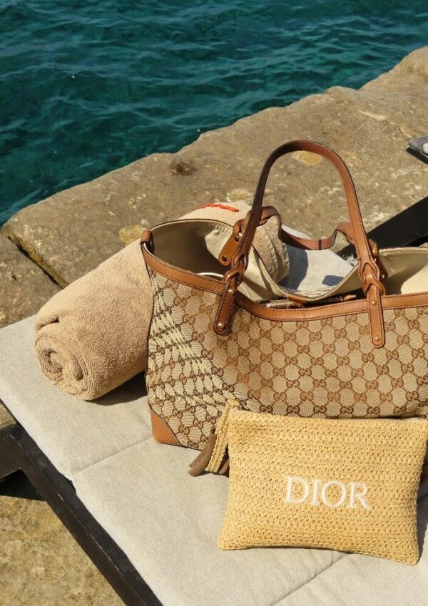 A rolled beige towel, a Gucci tote bag, and a woven Dior pouch rest on a lounge chair by a stone platform next to turquoise water.
