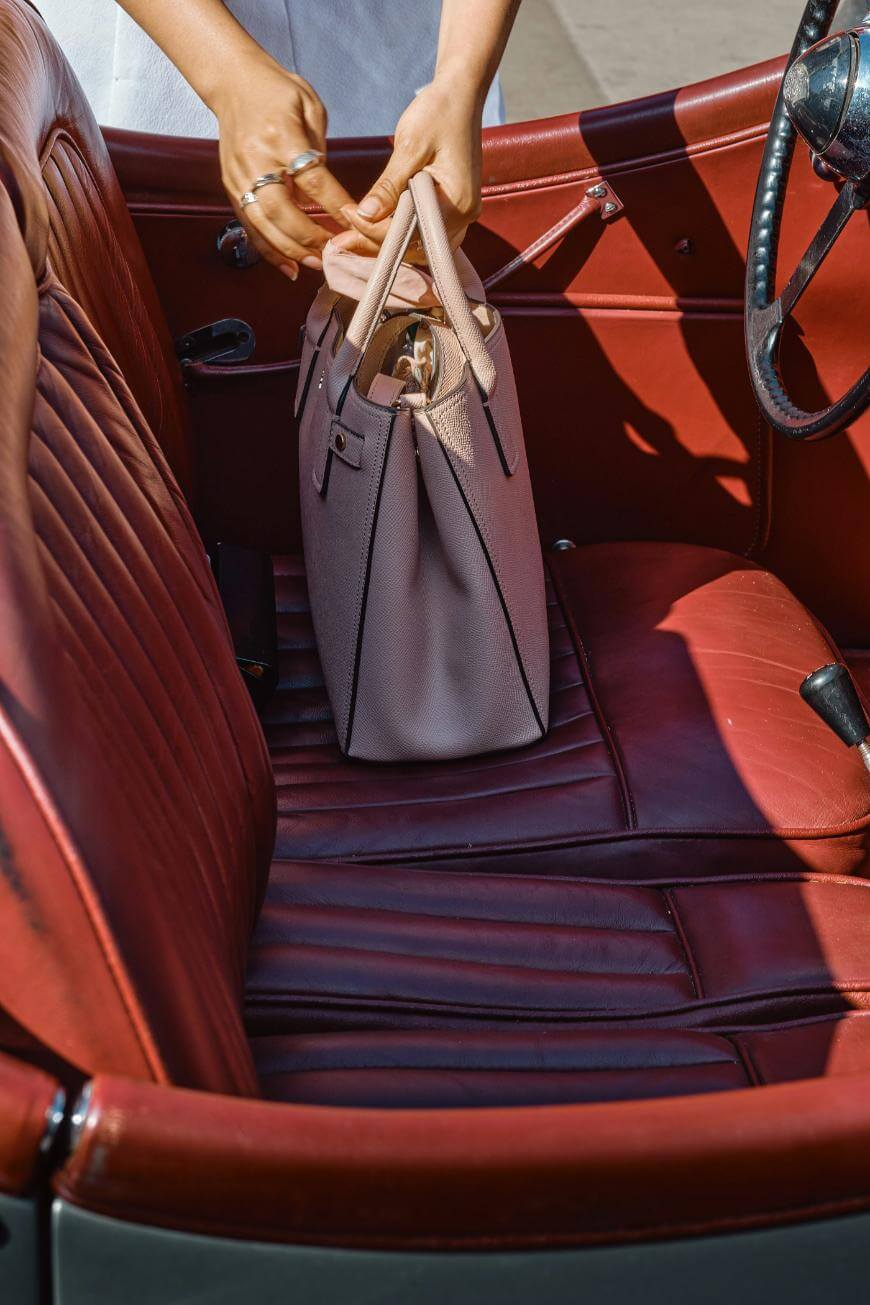 A person places a light pink handbag on the red leather seat of a vintage car, with one hand holding the bag and the other adjusting the handles.