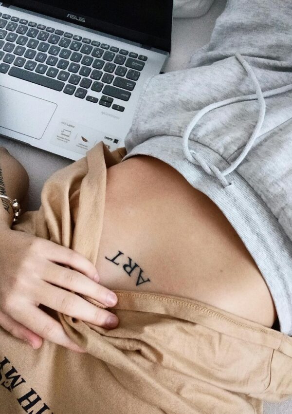 A person in grey sweatpants and a tan shirt lifts their shirt to reveal a tattoo that says ART on their ribcage. Part of a laptop is visible in the background.
