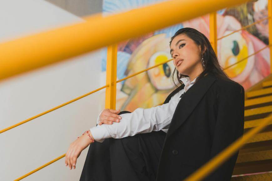 A woman in a black blazer and white shirt sits on yellow stairs, looking thoughtful. Colorful wall art is visible in the background.