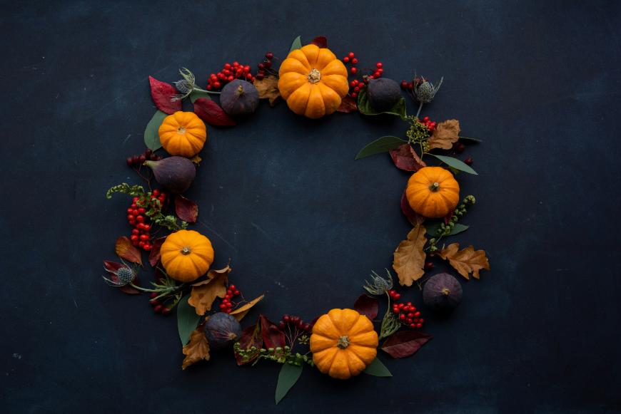 A circular autumn wreath made of small orange pumpkins, dark purple figs, red berries, and assorted fall leaves arranged on a dark background.