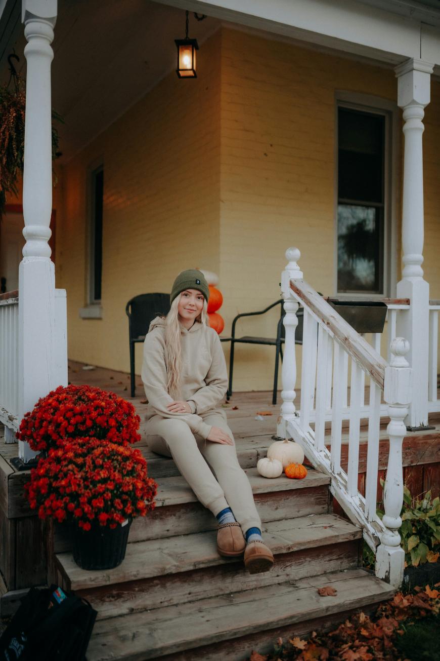 A woman in beige clothing and a green beanie sits on the steps of a porch decorated with red flowers and pumpkins. She is smiling, and the house behind her has yellow walls and white railings.