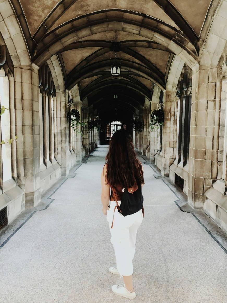 A person with long brown hair, wearing white pants and a black backpack, walks down a vaulted stone corridor with arched ceilings and hanging lanterns.