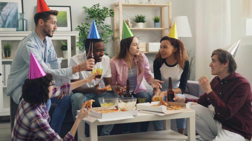 A group of six young adults wearing colorful party hats sit around a coffee table with pizza, popcorn, and drinks, chatting and laughing in a brightly lit living room decorated with plants and shelves.