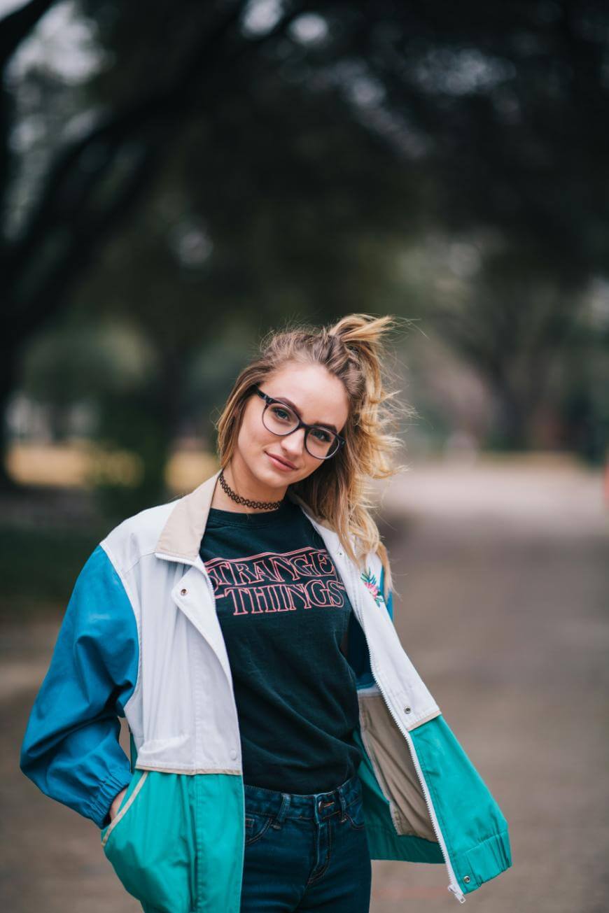 A young woman with long blonde hair, glasses, and a choker necklace stands outdoors on a path, wearing a retro jacket and a Stranger Things T-shirt, smiling slightly with her hands in her pockets.