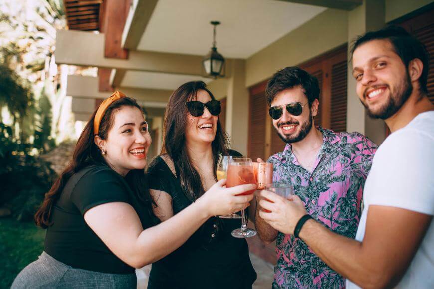Four friends stand outside, smiling and clinking their drinks together in a cheerful toast. They appear relaxed and happy, enjoying a sunny day.