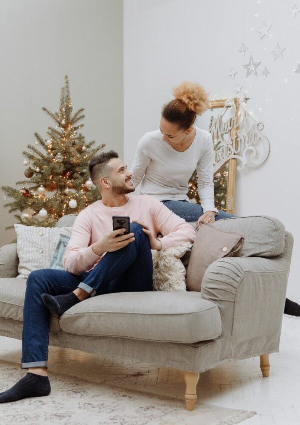 A man sitting on a beige sofa holding a phone looks up and smiles at a woman kneeling behind him, sharing one of their first holidays together. A decorated Christmas tree and festive ornaments are in the background.