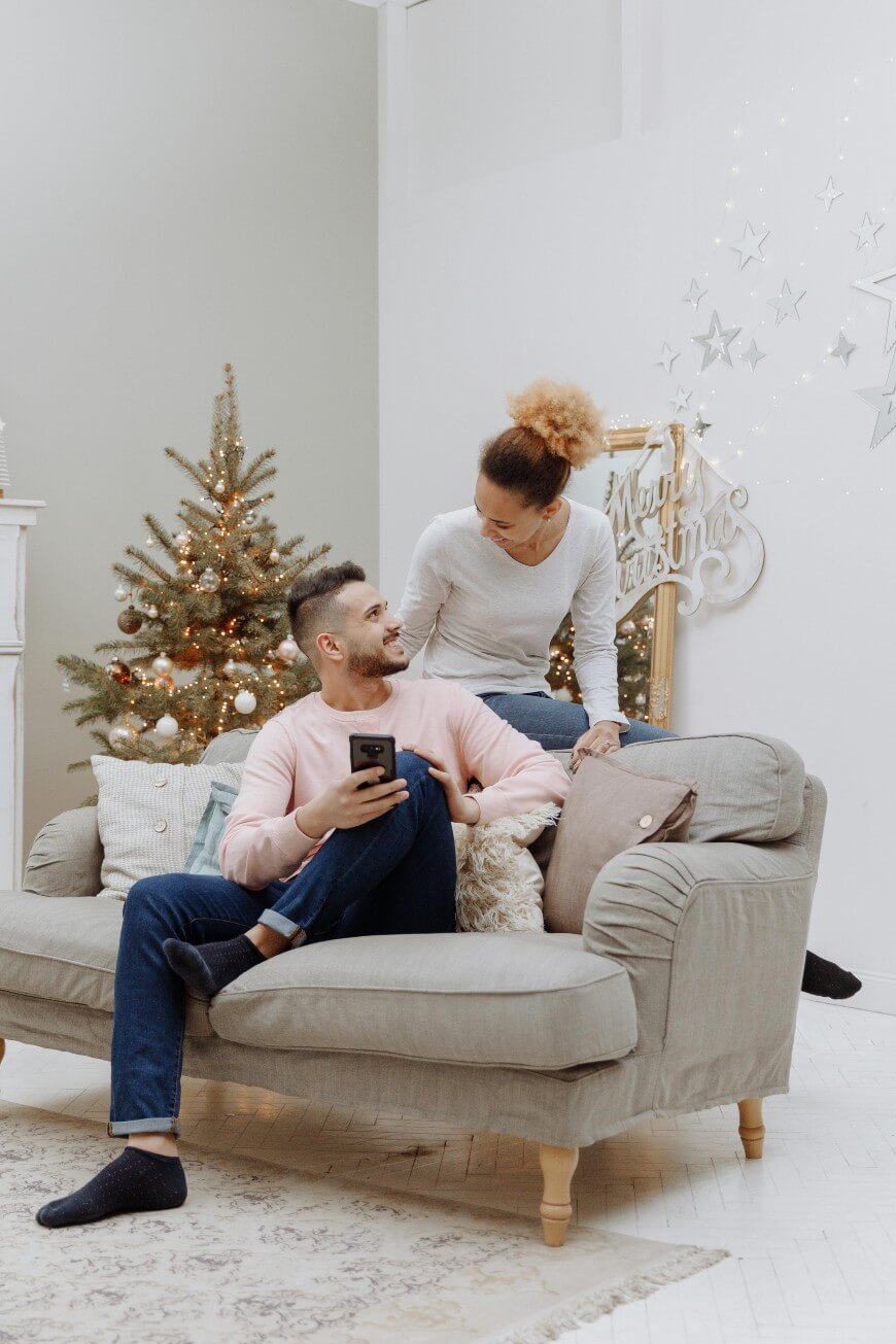 A couple enjoys their first holidays together in a cozy living room, decorated for Christmas with a tree and festive lights. One person sits on the sofa with a phone, while the other stands behind, smiling and leaning on the sofa’s back.