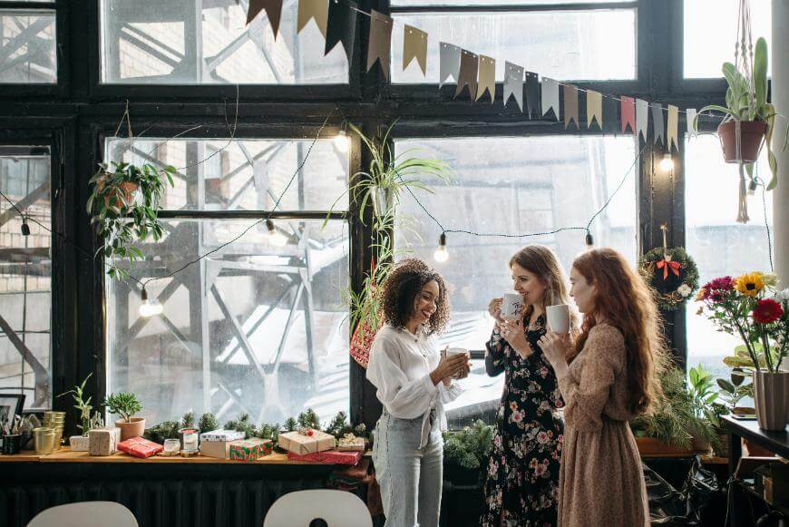 Three women stand together indoors by large windows, holding mugs and smiling, surrounded by potted plants, string lights, flowers, and colorful bunting decorations. Wrapped gifts are visible on the table beside them.