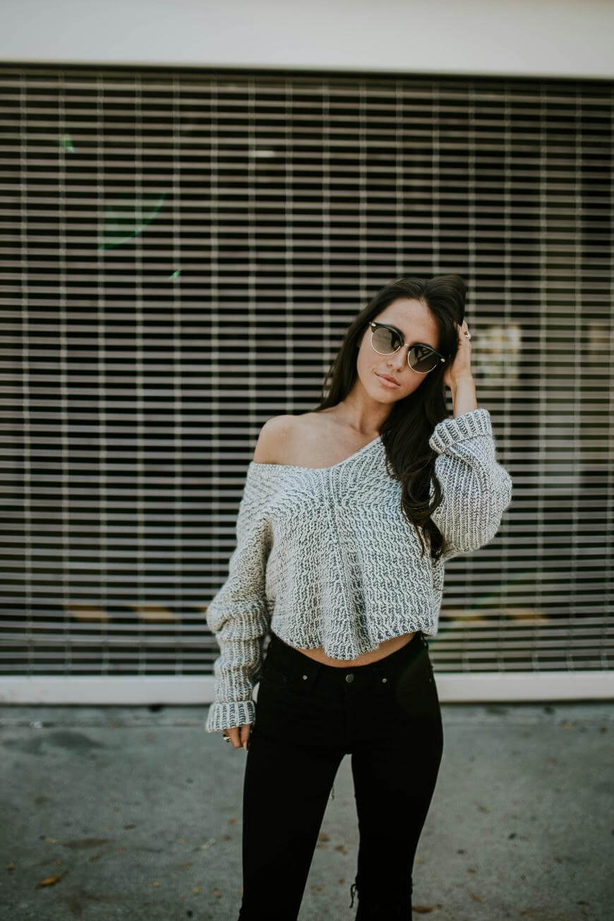 A woman wearing sunglasses, an off-the-shoulder knitted sweater, and black pants stands in front of a metal security gate, with one hand touching her hair and looking at the camera.