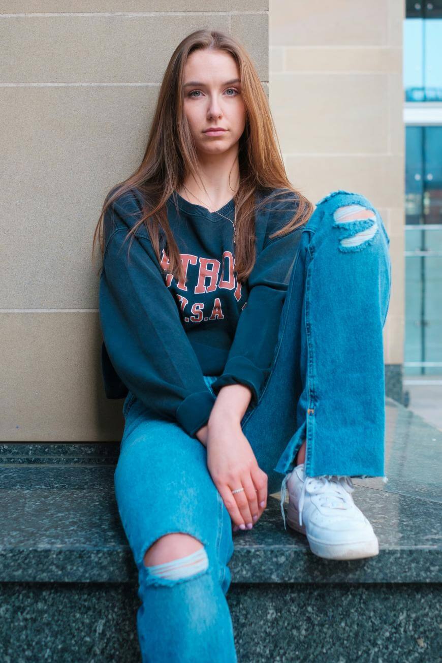A young woman with long brown hair sits on a stone bench, wearing a dark sweatshirt with PATBOY USA and ripped blue jeans. She looks at the camera with a neutral expression, one knee raised, and white sneakers.