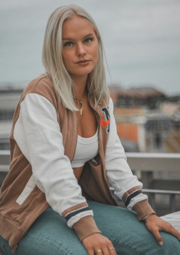 A young woman with straight blond hair wearing a tan and white jacket, a white crop top, and blue jeans sits outdoors on a metal railing, looking directly at the camera with a neutral expression.