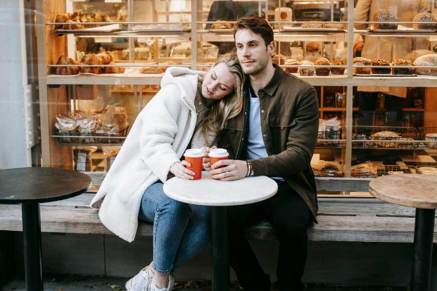 A woman leans her head on a mans shoulder as they sit together at an outdoor café table, each holding a cup. Behind them is a bakery window filled with pastries and breads.