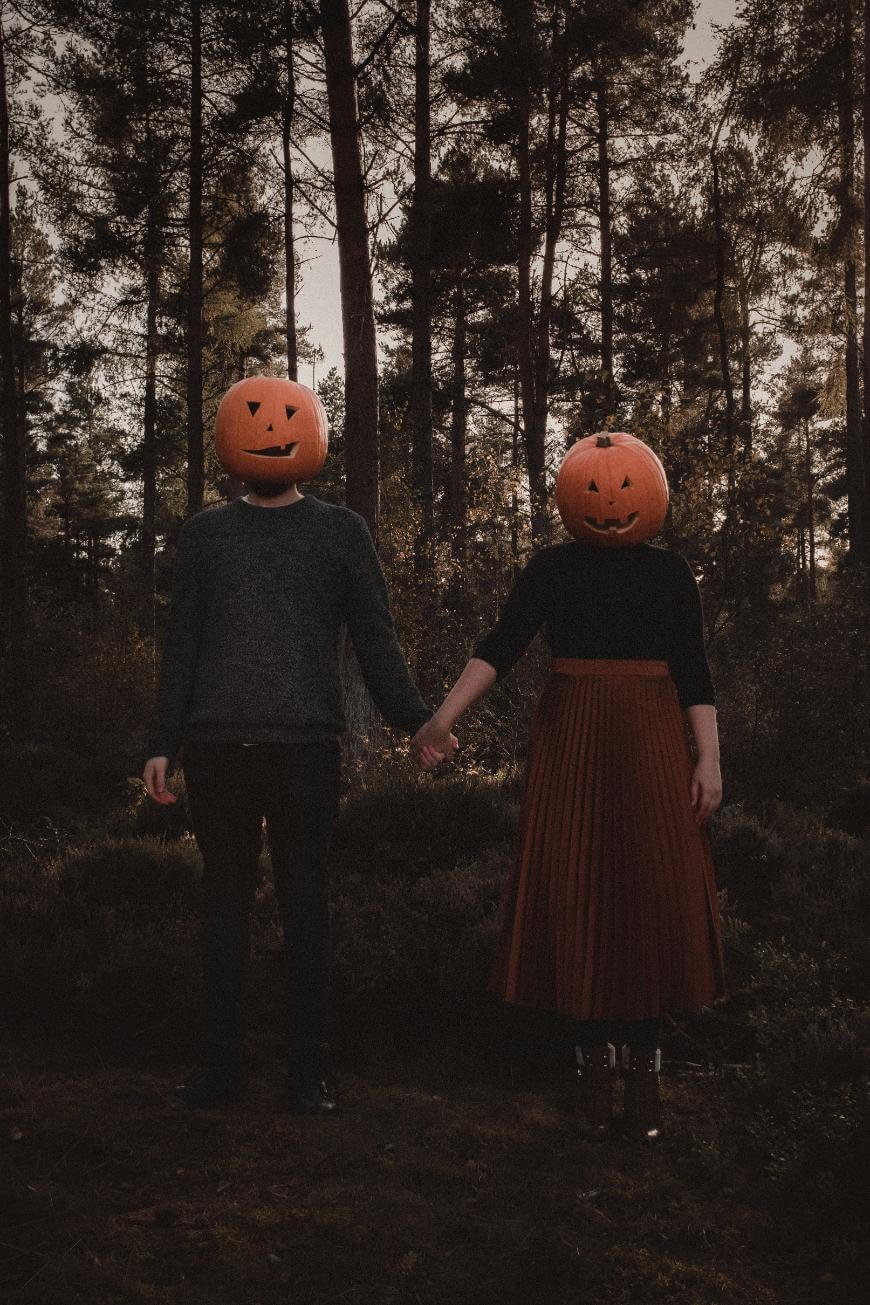 Two people stand hand in hand in a forest, each wearing a carved pumpkin as a mask. The scene is dimly lit, creating a spooky, autumn atmosphere.