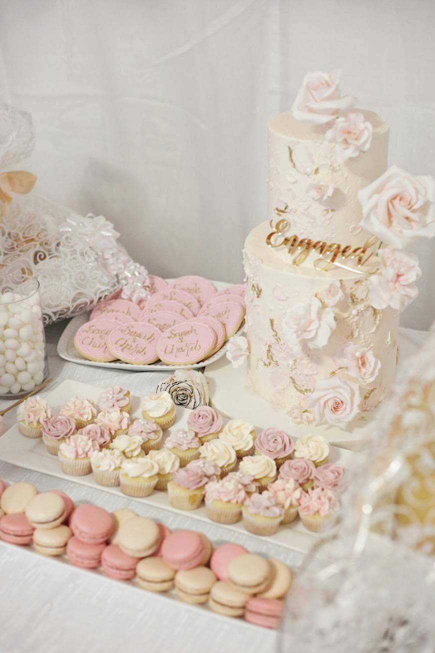 A table display with a white and pink engagement cake decorated with flowers, pink cookies reading Engaged, mini cupcakes with pastel frosting, assorted macarons, and candies in glass jars.
