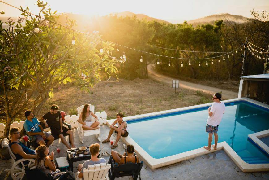 A group of friends gathers by a swimming pool at sunset, sitting and chatting on chairs, while one person stands on the pool’s edge. String lights hang above, and hills and trees are seen in the background.