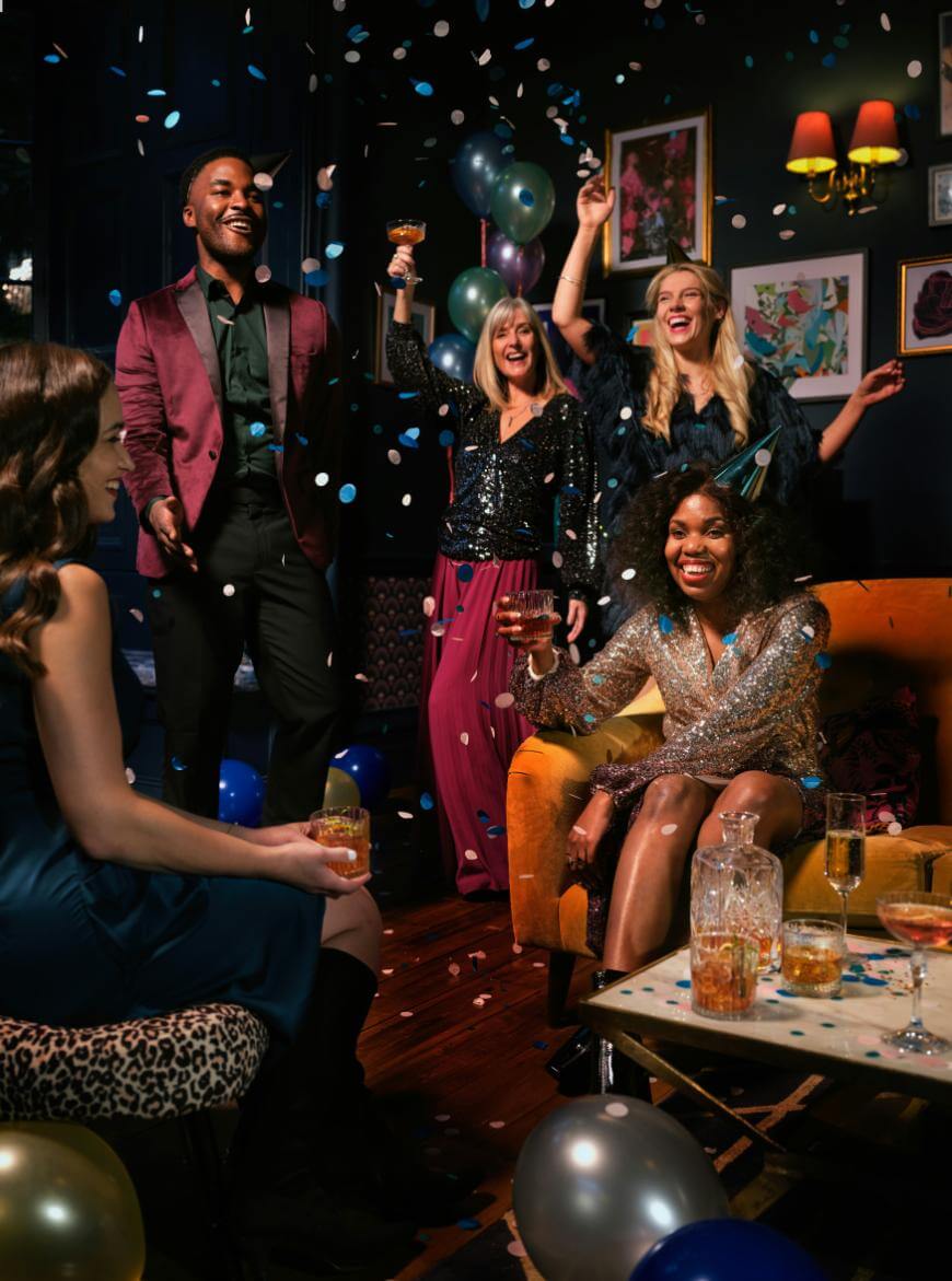 A group of five people celebrate at a lively party indoors, smiling and throwing confetti. They are dressed in festive outfits, with drinks in hand. Balloons and decorations fill the room, creating a joyous atmosphere.