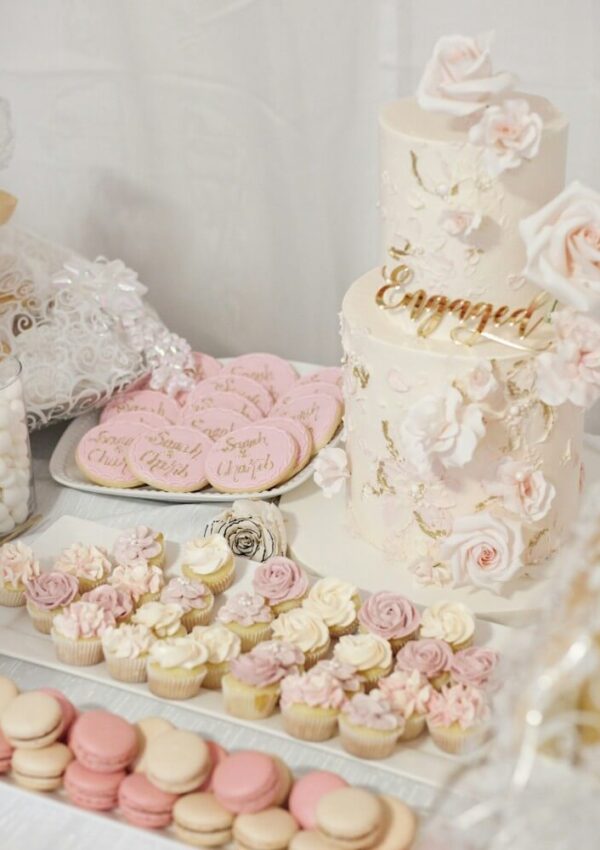 A dessert table displays a two-tiered white and pink floral cake with a gold Engaged topper, pink decorated cookies, flower-topped cupcakes, and pink and cream macarons.