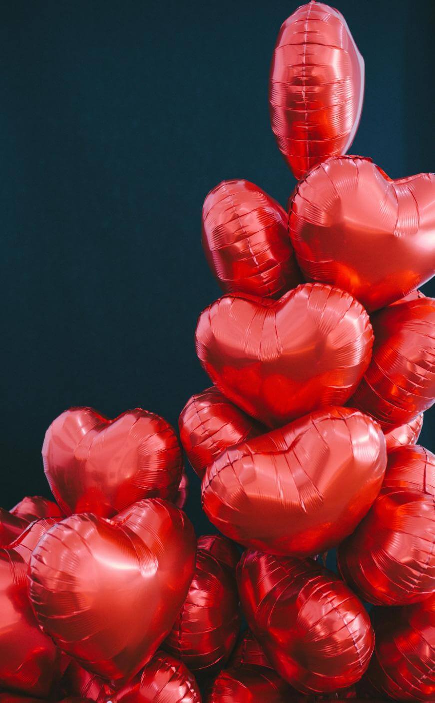 A bunch of shiny red heart-shaped balloons are stacked together against a dark background.