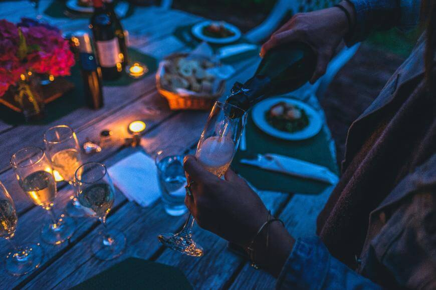 A person pours sparkling wine into a glass at an outdoor dinner table set with candles, food, and flowers during the evening.