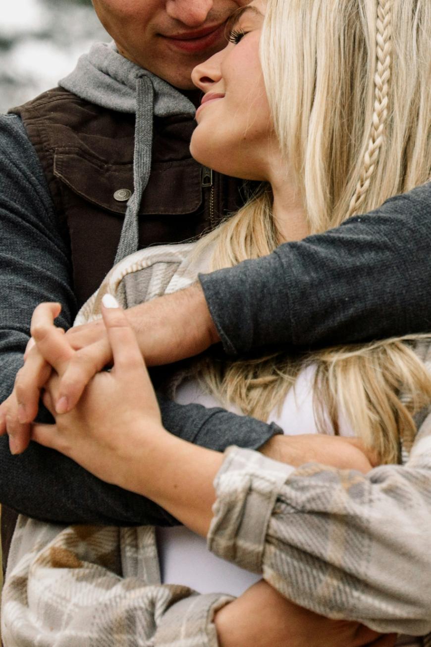 A close-up of a couple embracing outdoors, with the man’s arms wrapped around the woman from behind as she smiles and leans into him. They are holding hands, and both wear casual, layered clothes.