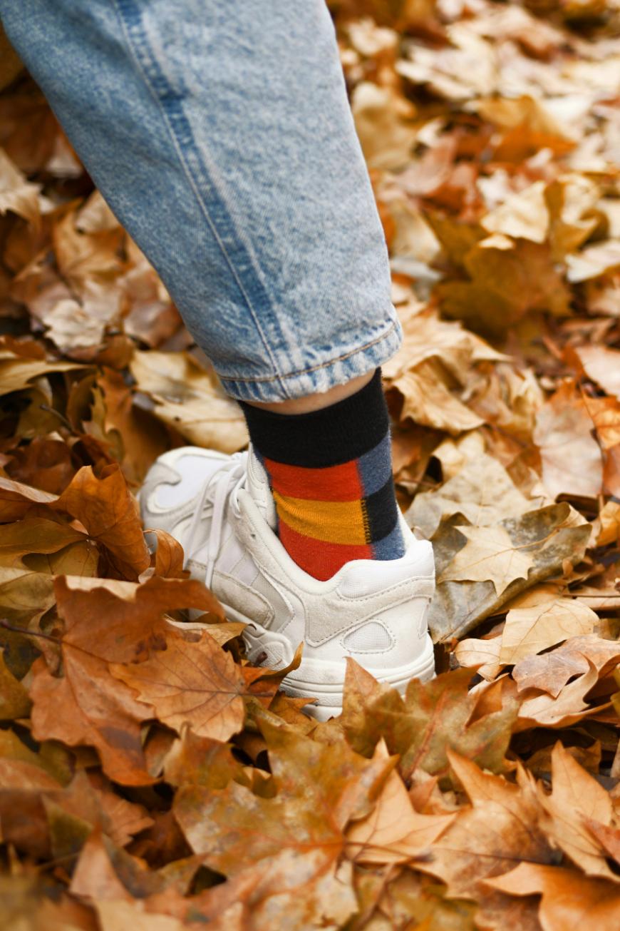 A person wearing rolled-up blue jeans, white sneakers, and colorful striped socks stands on dry autumn leaves scattered on the ground.