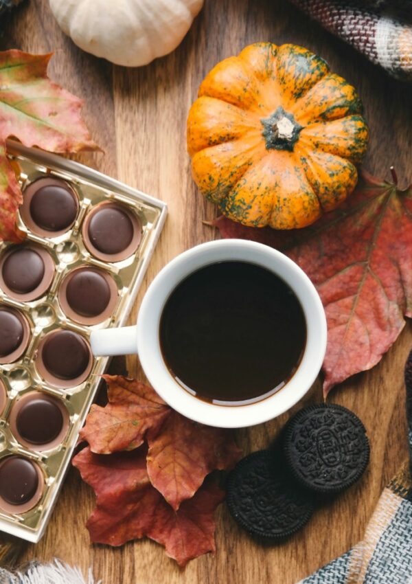 A white cup of black coffee sits on a wooden table surrounded by autumn leaves, a small pumpkin, assorted chocolates, and chocolate sandwich cookies, with a plaid scarf partially visible in the corner.
