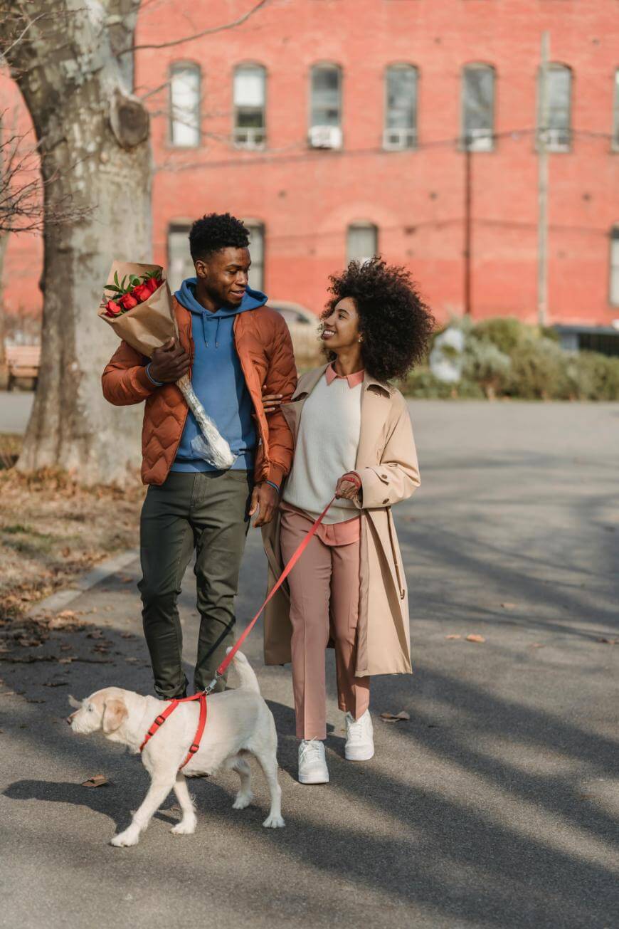 A man holding a bouquet of flowers walks hand-in-hand with a woman, both smiling at each other, while she leads a dog on a red leash down a city sidewalk. A red brick building is in the background.