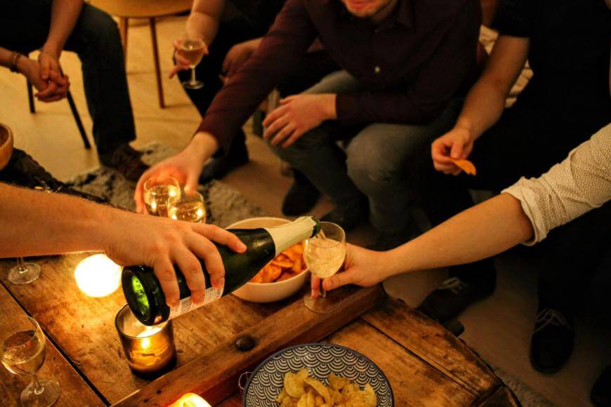 A group of people sit around a wooden table with snacks and lit candles, while one person pours sparkling wine into another’s glass.