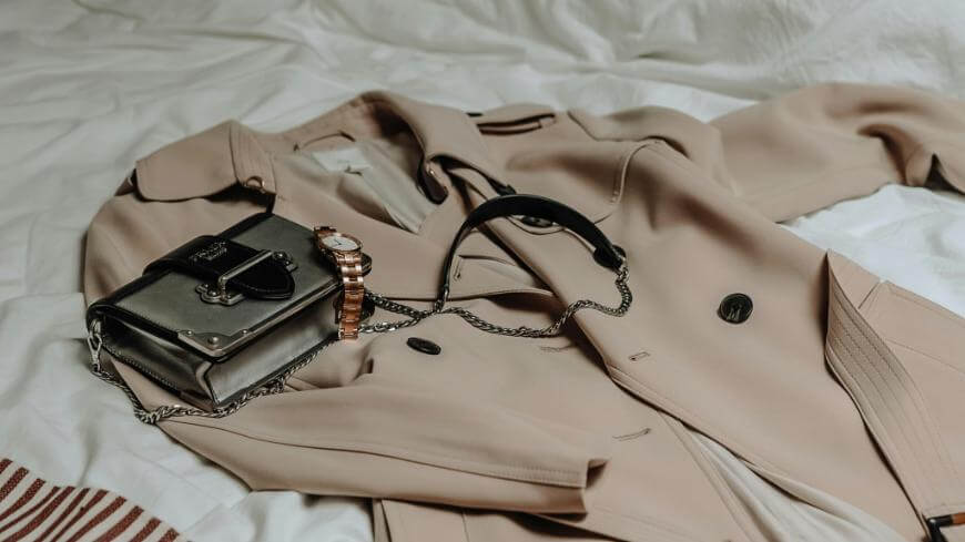 A beige trench coat, a black handbag with a chain strap, and a rose gold wristwatch are neatly arranged on a white bedspread.