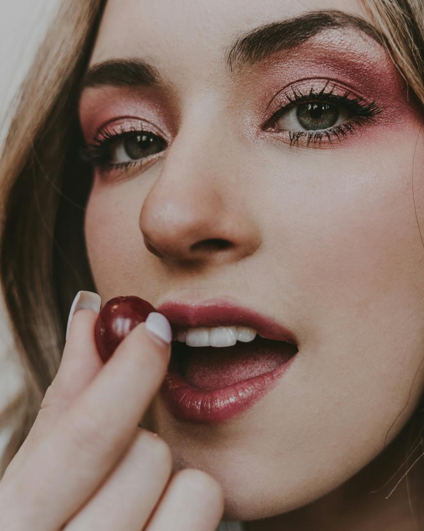 A close-up of a person with pink eyeshadow and lip color, holding a cherry near their slightly open lips, with manicured nails visible.