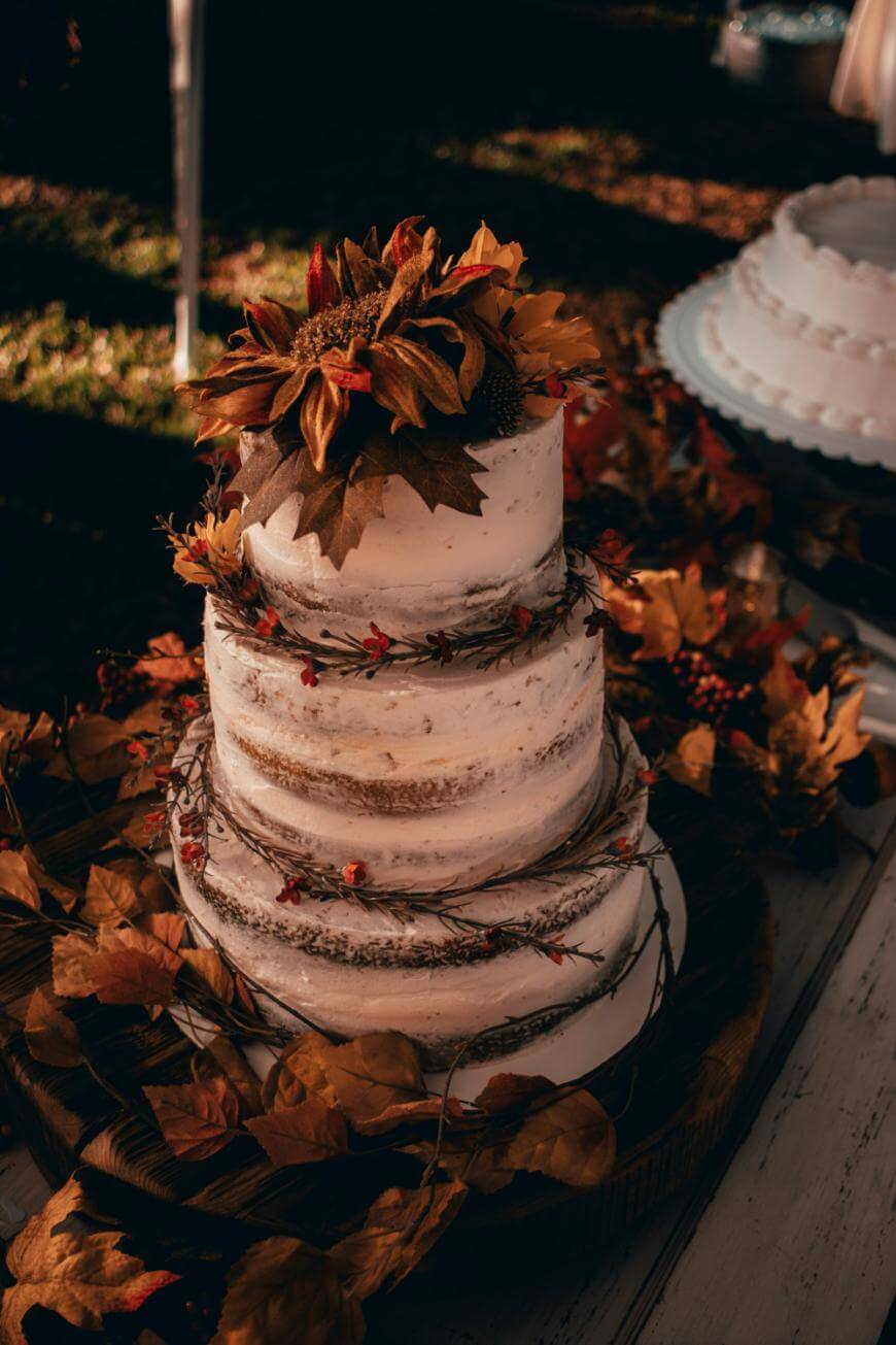 A three-tiered rustic naked cake decorated with twigs, berries, and autumn leaves, topped with large artificial sunflowers. The cake sits on a wooden platter, surrounded by fall foliage.
