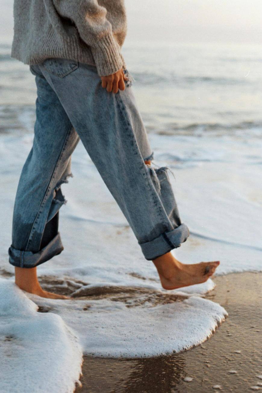 A person wearing rolled-up jeans and a sweater walks barefoot along the shoreline, letting gentle waves and sea foam touch their feet. The scene evokes a calm, relaxed mood at the beach.