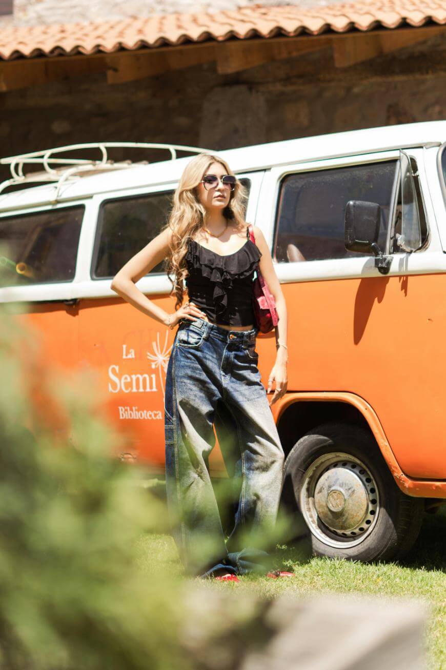 A woman wearing sunglasses, a black ruffled top, and loose-fitting jeans poses confidently in front of an orange and white van on a sunny day. Grass and a building with a tiled roof are visible in the background.