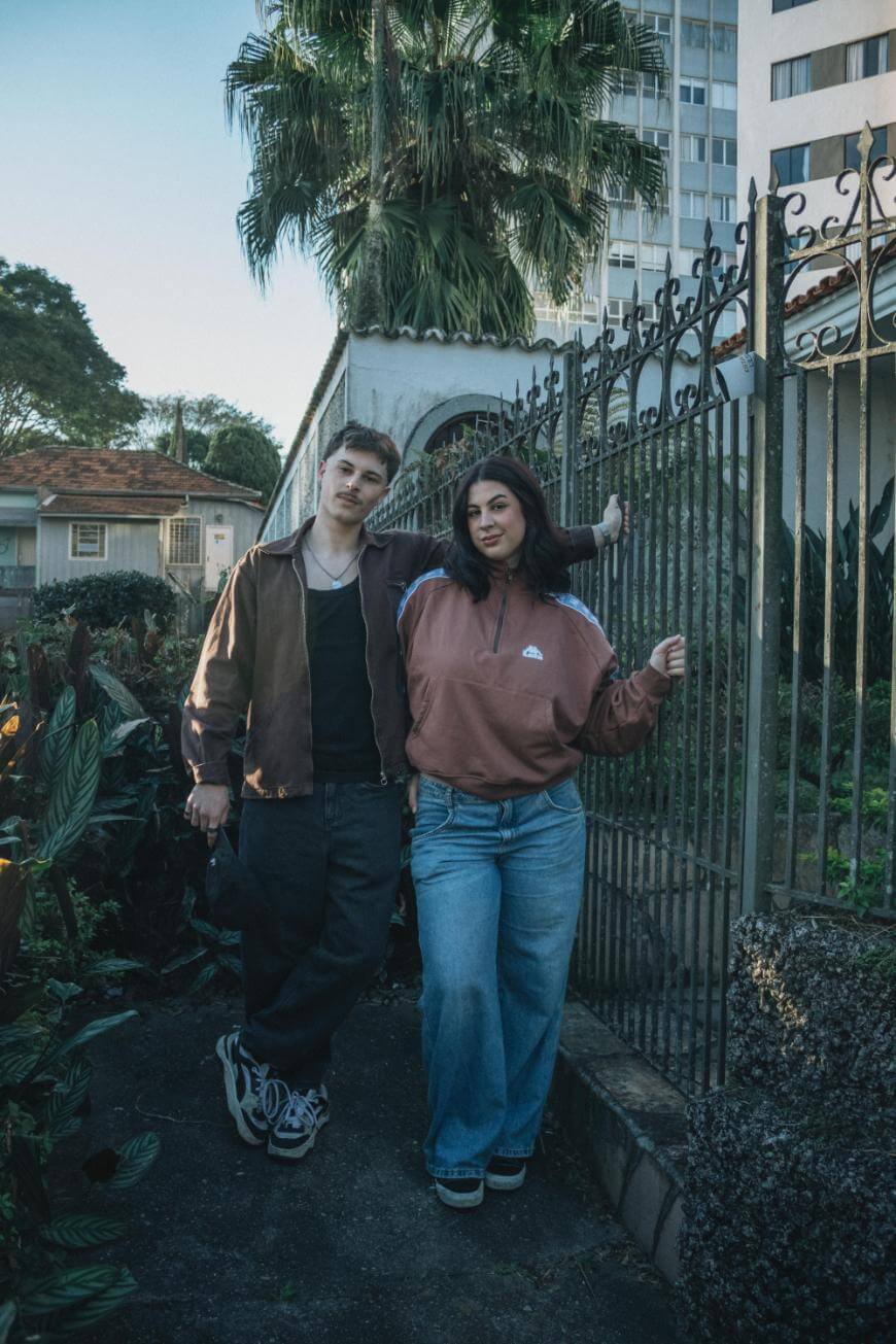 Two young adults stand side by side by a metal gate, surrounded by greenery. Both wear casual loose-fitting clothes. A tall palm tree and a multi-story building are visible in the background.