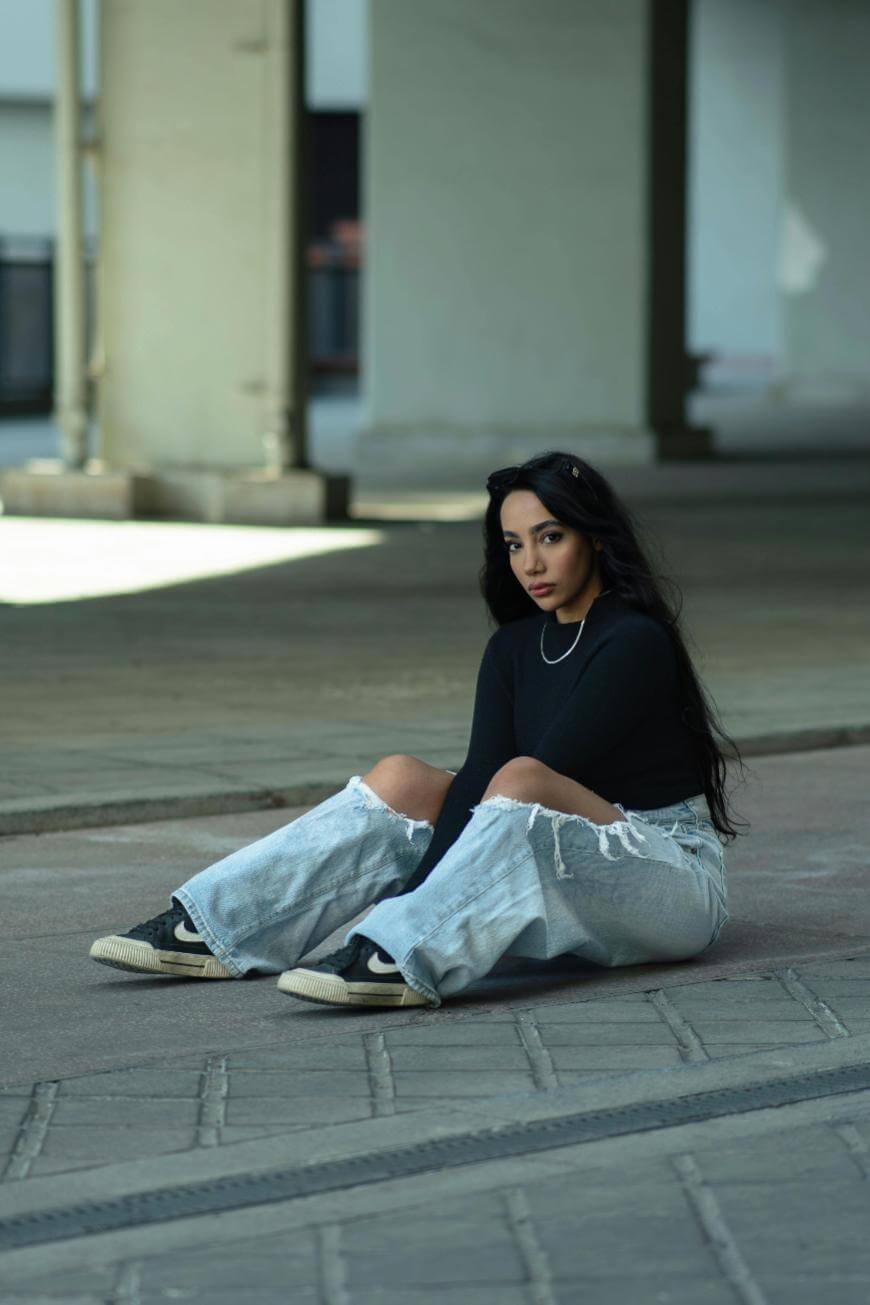 A woman with long dark hair sits on the ground in an urban area, wearing a black long-sleeve top, ripped light blue jeans, and black sneakers, looking directly at the camera with a neutral expression.