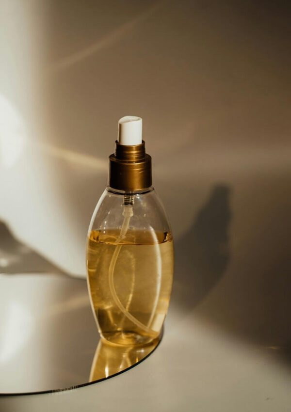 A clear spray bottle filled with golden liquid, featuring a white nozzle and gold-colored cap, stands on a reflective surface with soft, warm lighting and subtle shadows in the background.