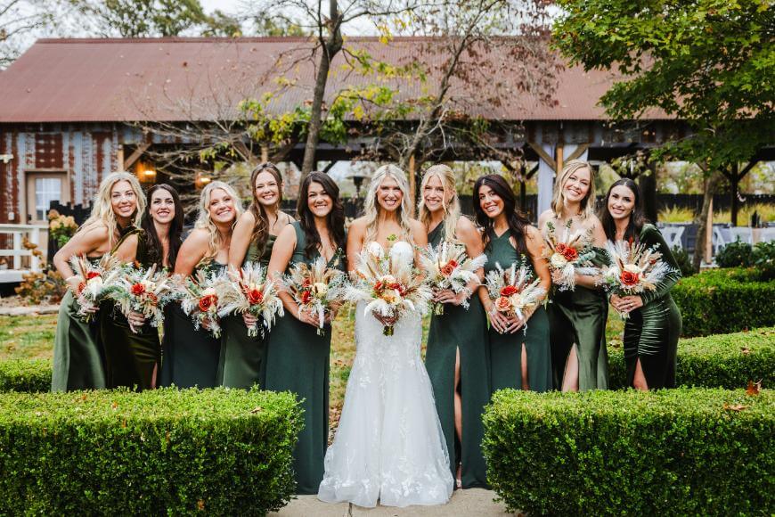 A bride in a white gown stands outside, smiling with ten bridesmaids in dark green dresses, each holding bouquets, with greenery, hedges, and a rustic barn in the background.