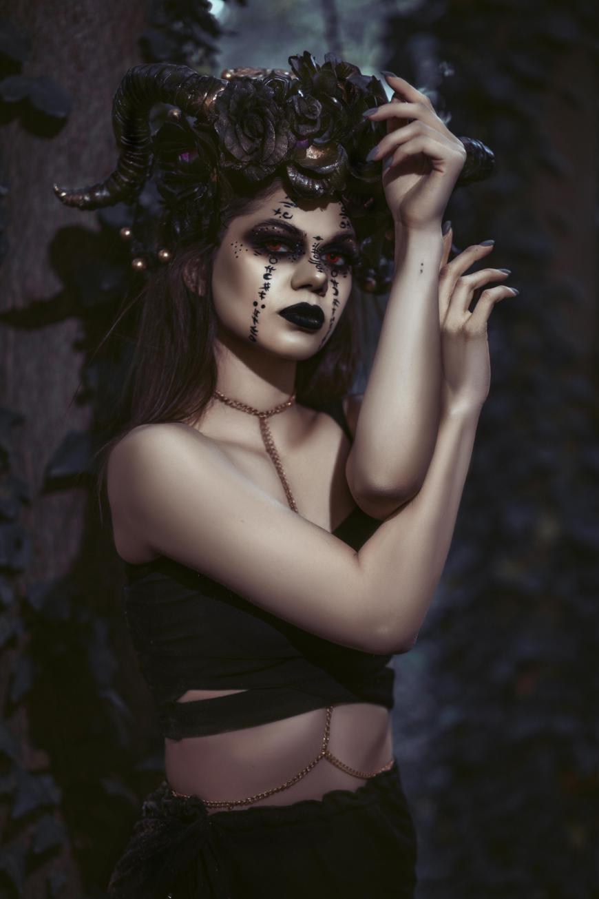 A woman in dark, dramatic makeup and black clothing poses in a forest, wearing a headpiece with large curled horns and black flowers. Her expression is intense, with hands raised near her face and adorned with gold chain jewelry.