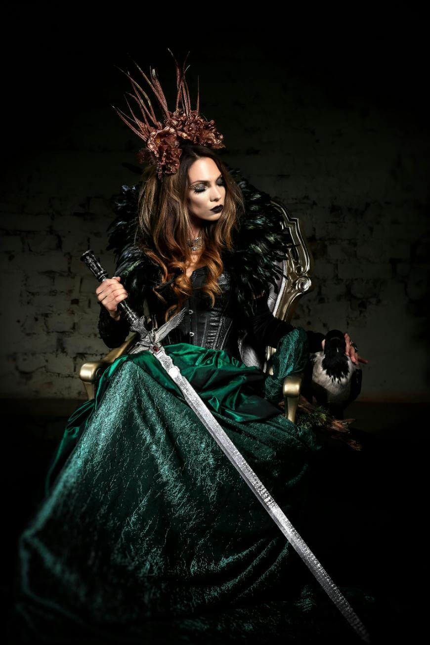 A woman in a dark feathered cape and green gown sits on a throne, holding a long sword. She wears dramatic makeup and a crown of twigs, exuding a powerful, fantasy queen aura against a shadowy background.