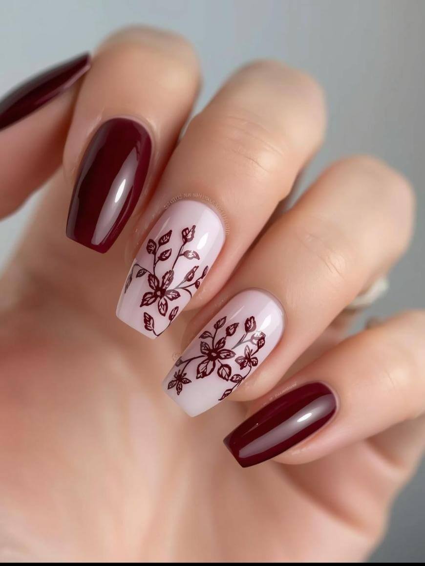 A hand with manicured nails; thumb, pinky, and index nails are glossy dark burgundy, while the middle and ring nails are pale pink with detailed burgundy floral designs.