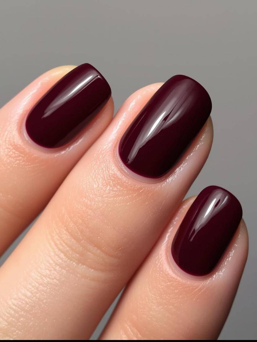 Close-up of three fingers with short, neatly manicured nails painted in a glossy, dark burgundy polish against a neutral background.