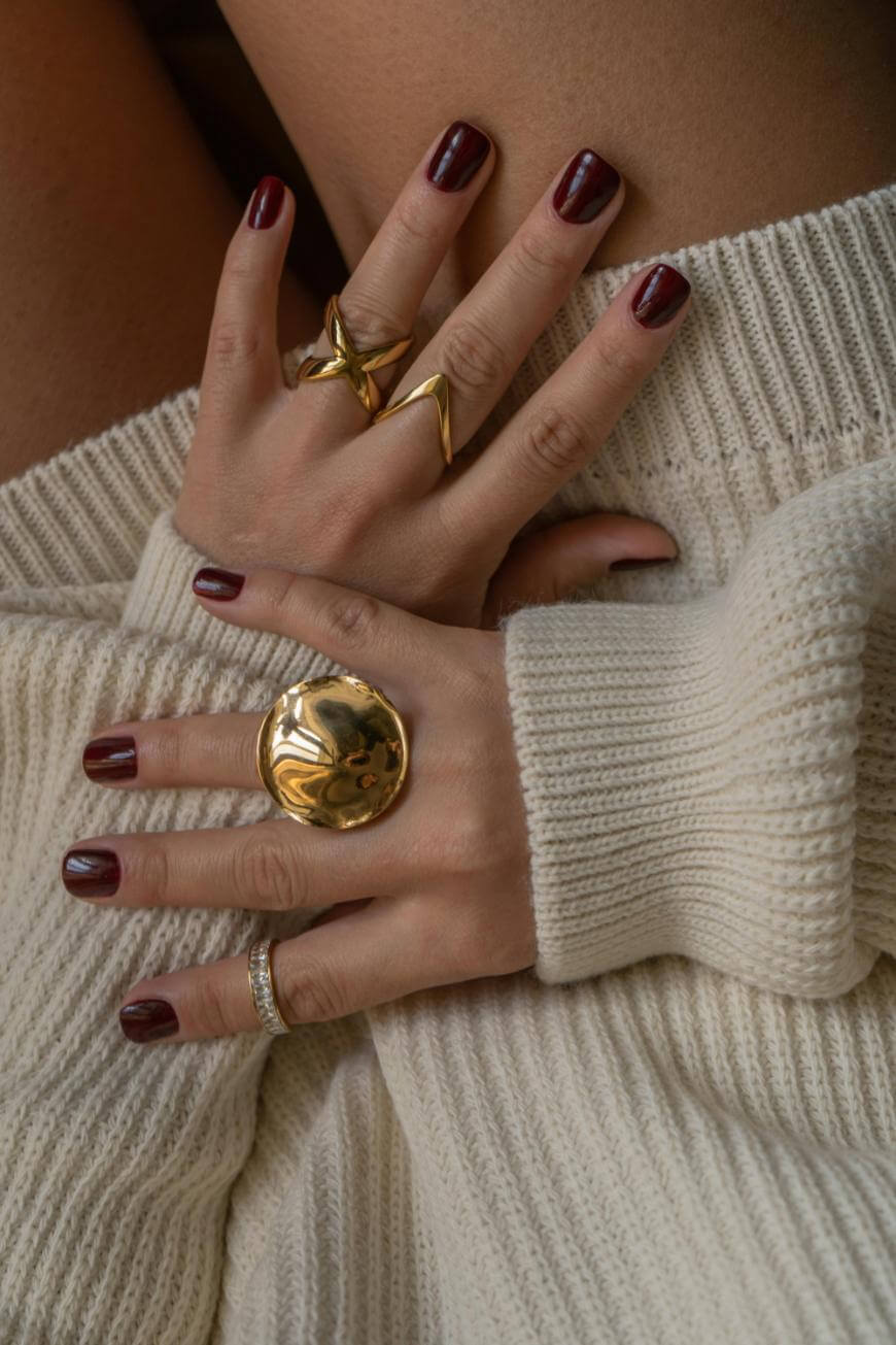 Close-up of two hands with dark red nail polish, wearing several gold and silver rings, resting on a cream-colored knit sweater.