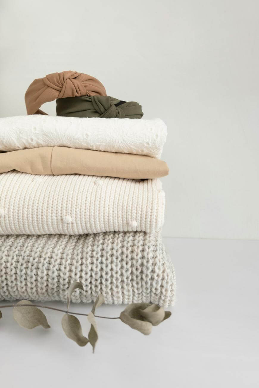 A neatly folded stack of neutral-colored clothes, including textured knits and a dotted fabric, with two headbands on top. Dried leaves are placed beside the stack against a plain white background.