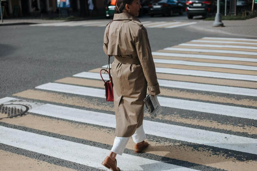 A person wearing a tan trench coat, white pants, and brown shoes crosses a city street at a crosswalk, holding a newspaper and carrying a small red shoulder bag.