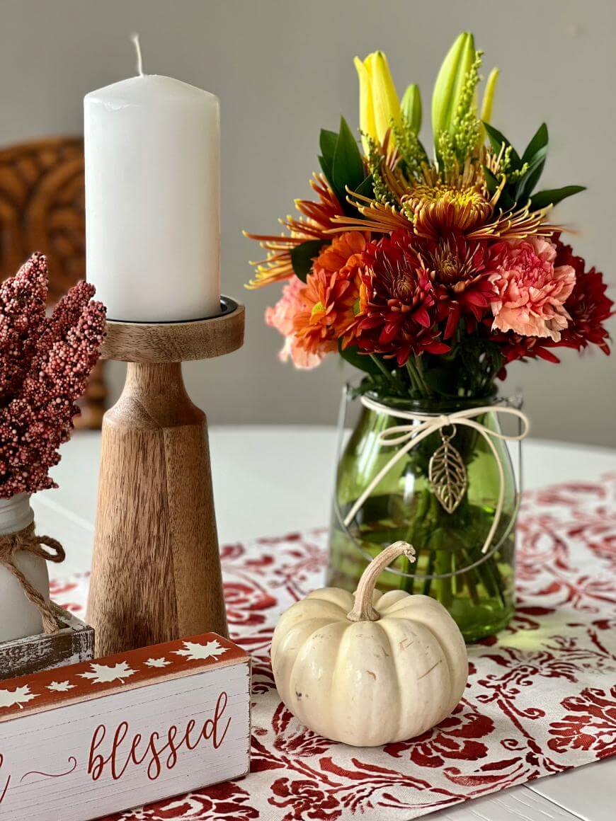 A fall-themed table centerpiece featuring a white candle on a wooden holder, a bouquet of orange and yellow flowers in a jar, a small white pumpkin, and a “blessed” sign, all on a red and white patterned table runner.