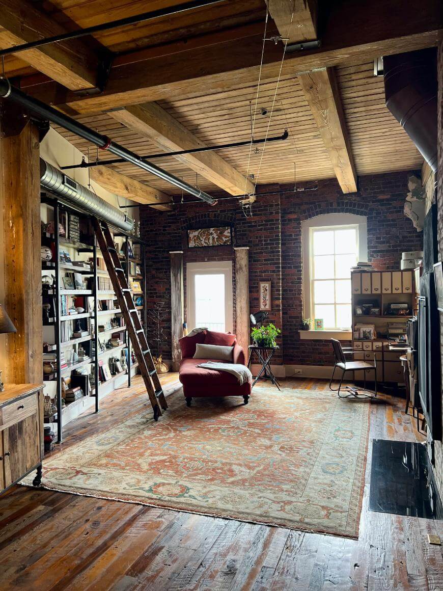 A cozy loft room with exposed brick walls, wooden beams, a large rug, a red armchair with an ottoman, a tall bookshelf with a ladder, a desk by the window, and various books and plants.