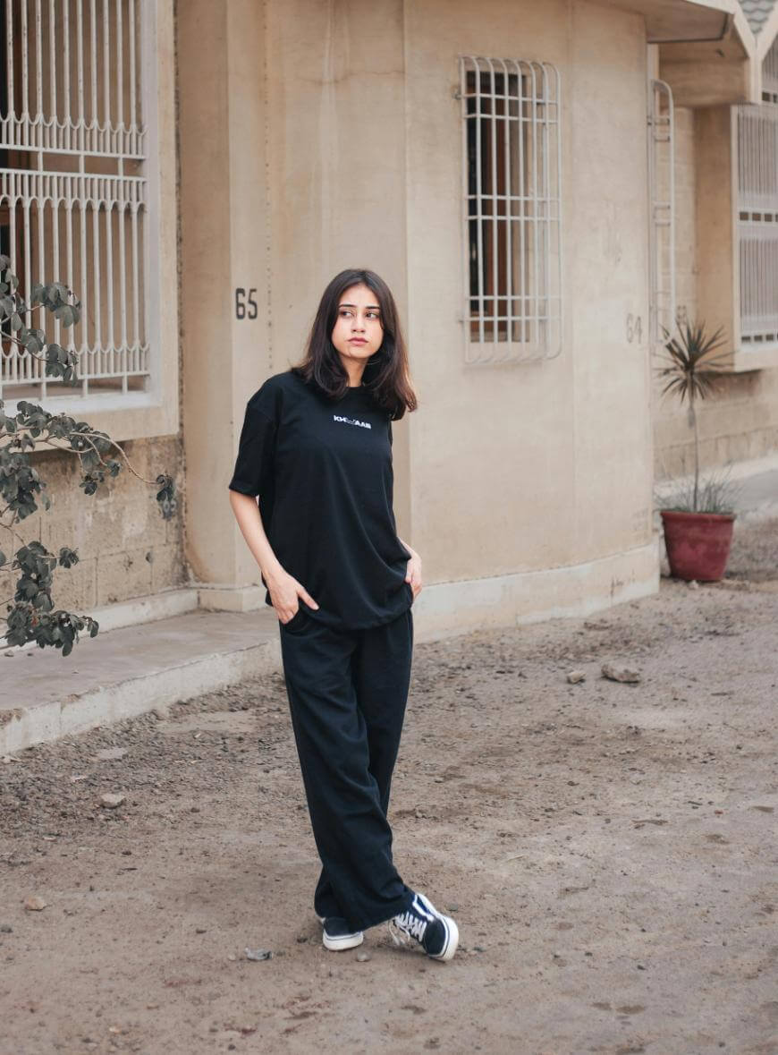 A woman with medium-length dark hair, wearing a black t-shirt, black pants, and sneakers, stands with hands in pockets on a dirt path near a beige building with barred windows and a potted plant.