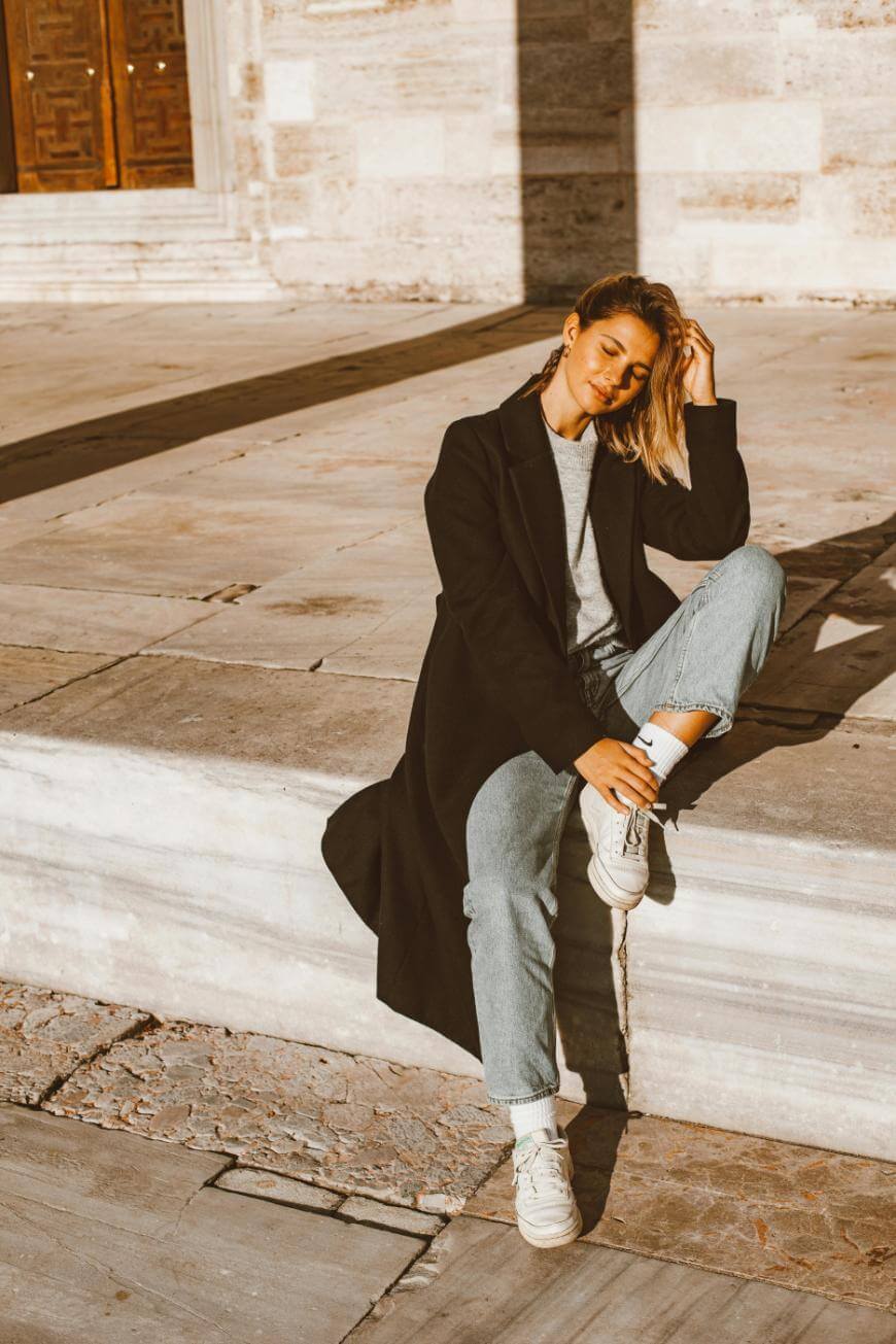A woman wearing a long black coat, jeans, and white sneakers sits on a stone ledge outdoors, smiling with one hand in her hair in warm sunlight. The background features light stone walls and wooden doors.