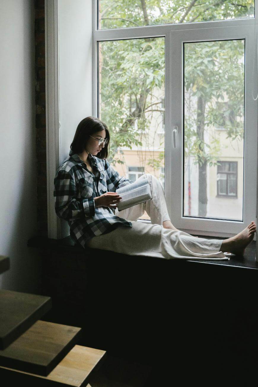 A woman wearing glasses and a plaid shirt sits on a windowsill, reading a book. Large windows behind her reveal green trees outside. She appears relaxed and comfortable in the natural light.